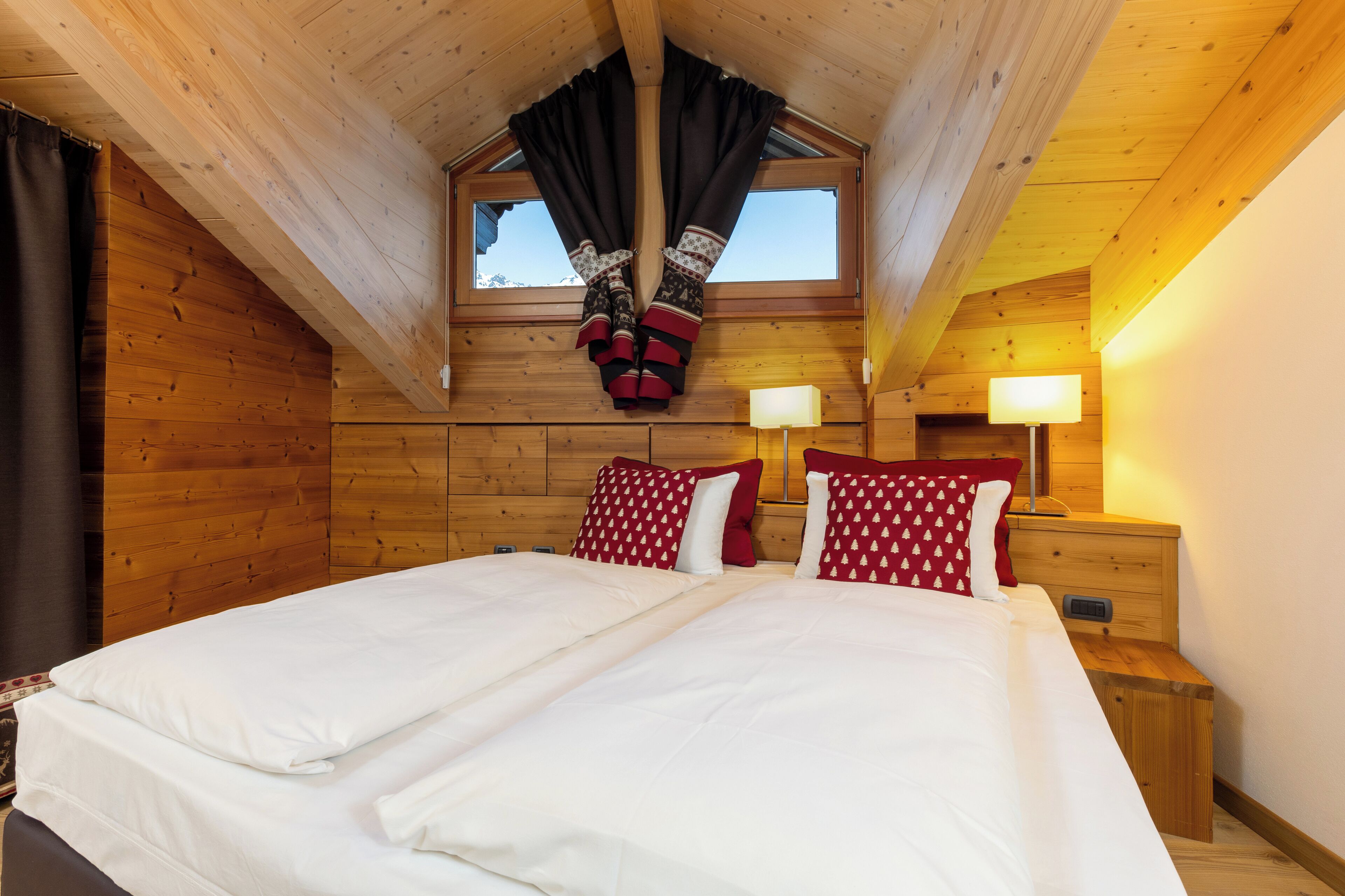 A bedroom with a wooden ceiling, a large bed with white bedding, two red pillows, and a window with curtains.