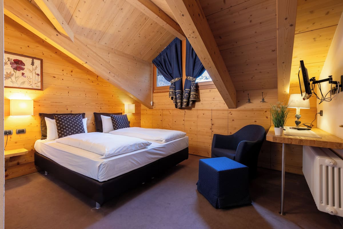 A cozy room with a wooden ceiling, a bed with white linens, a desk with a lamp, and a window with curtains.