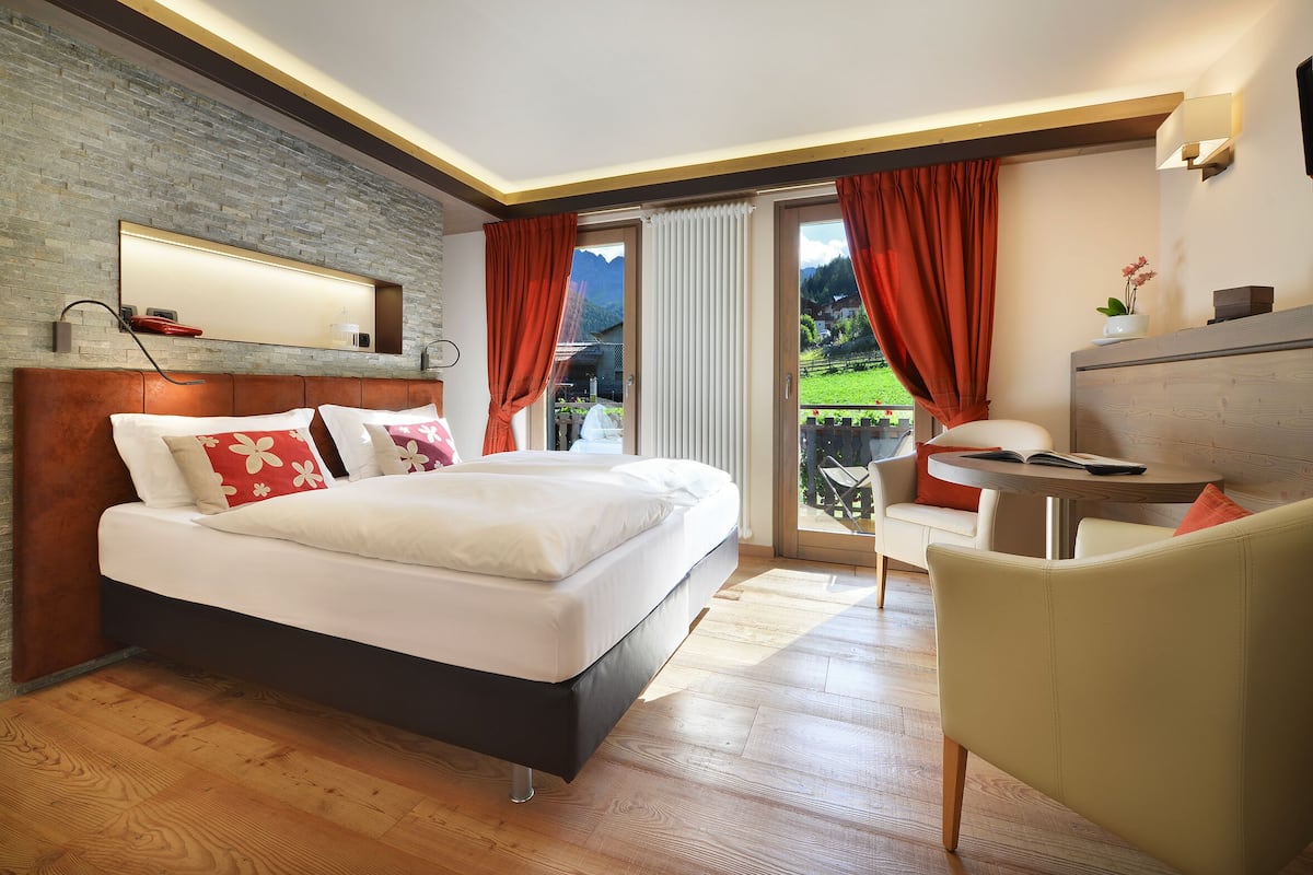 A modern hotel room with a large bed, a wooden floor, a balcony with red curtains, and a view of the mountains.