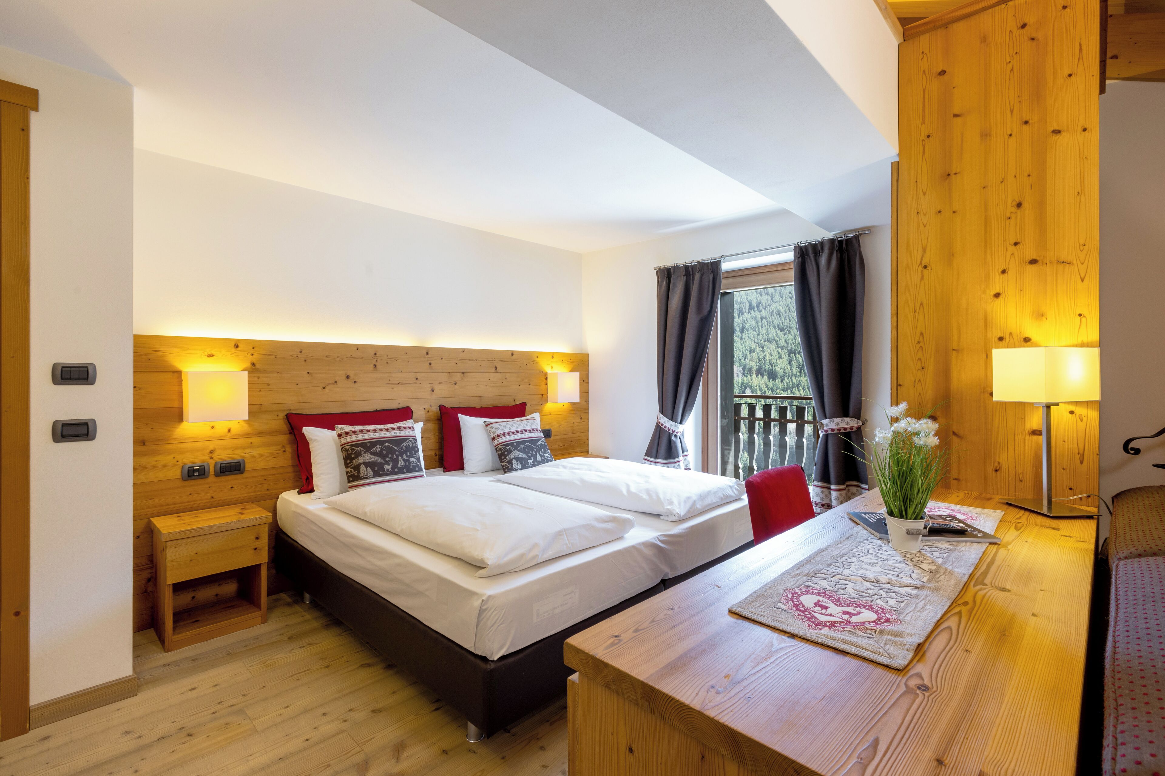Un dormitorio con cabecera de madera, una cama doble con ropa de cama blanca, una mesita de noche con lámpara y vistas al exterior a través de una ventana con cortinas.