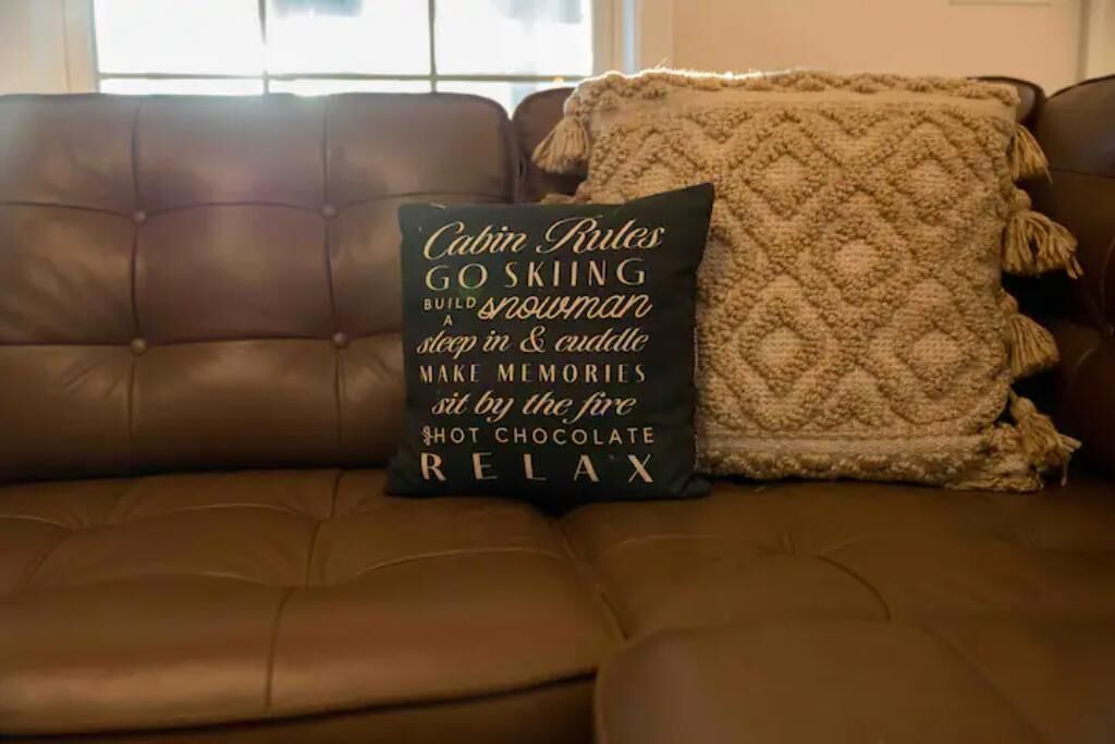 A brown leather couch with a cushion that reads "Cabin Rules" and includes phrases like "GO SKIING," "BUILD A SNOWMAN," "sleep in & cuddle," "MAKE MEMORIES," "sit by the fire," "SHOT CHOCOLATE," and "RELAX."