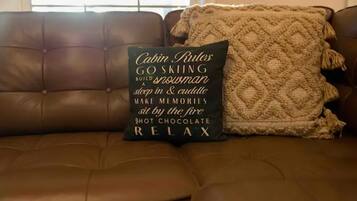 A brown leather couch with a cushion that reads "Cabin Rules" and includes phrases like "GO SKIING," "BUILD A SNOWMAN," "sleep in & cuddle," "MAKE MEMORIES," "sit by the fire," "SHOT CHOCOLATE," and "RELAX."