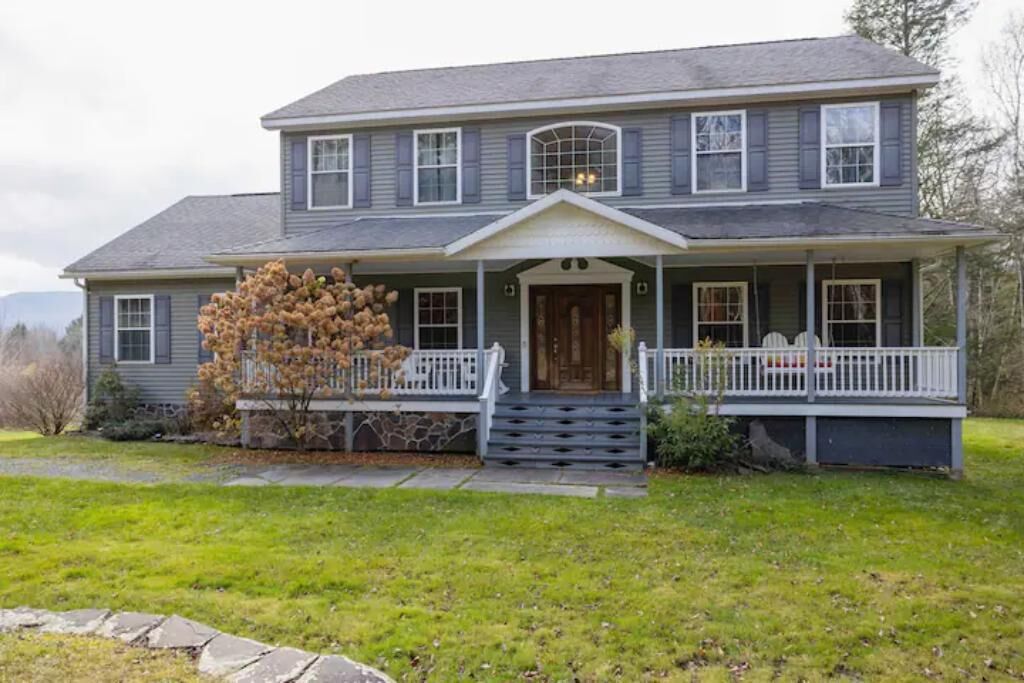 A two-story house with a front porch, a stone foundation, and a large tree in front.