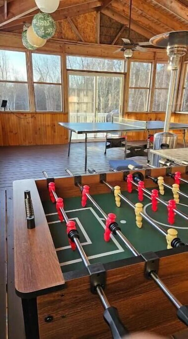 A room with a foosball table, a ping pong table, and a ceiling fan.