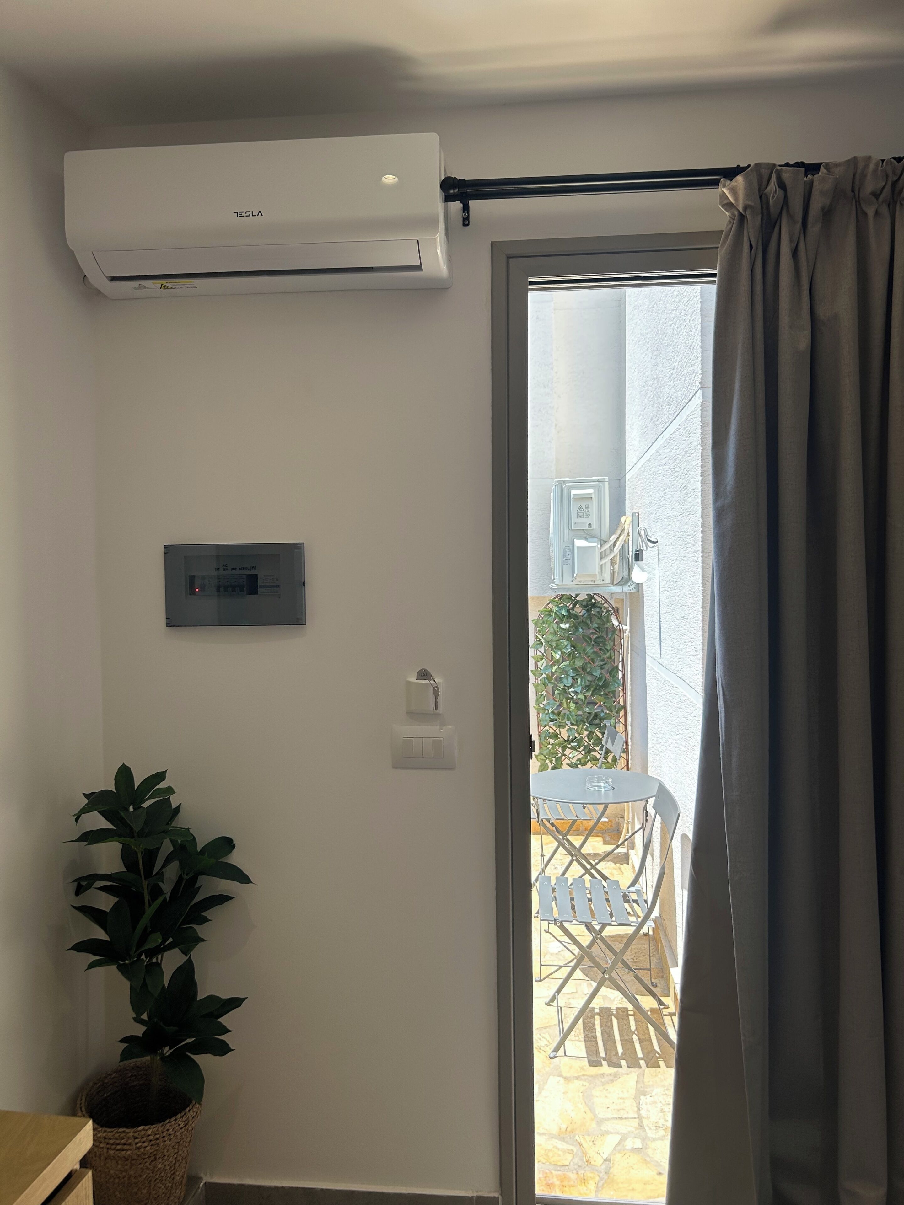 A room with a ceiling-mounted air conditioner, a wall-mounted control panel, a potted plant, and a view of an outdoor patio with a table and chairs.
