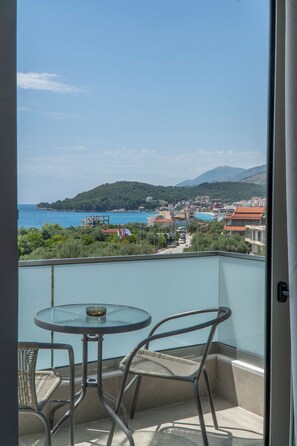 A balcony with a glass table and two wicker chairs, overlooking a coastal town with buildings and a mountain in the distance.
