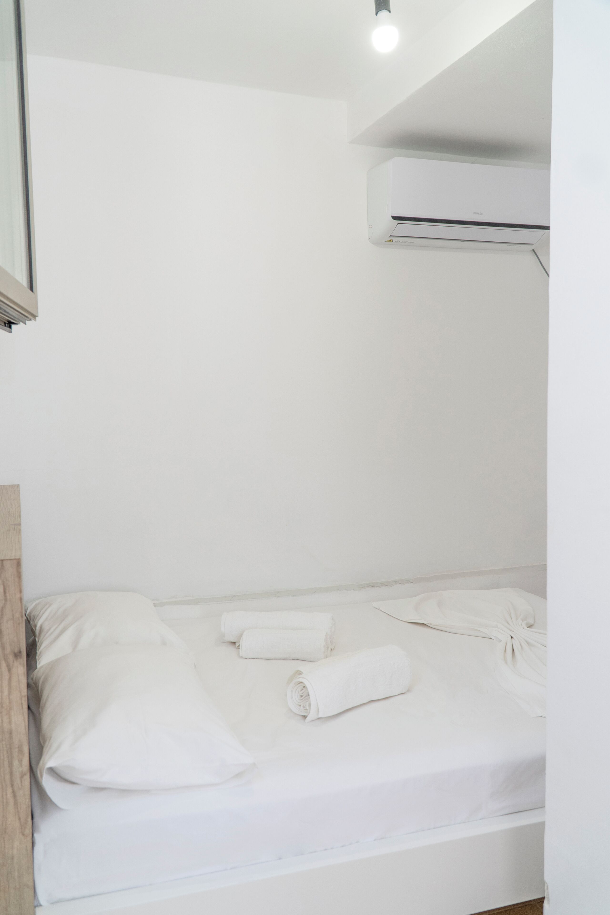 A single bed with white linens and a rolled towel, a wooden headboard, and a wall-mounted air conditioning unit.