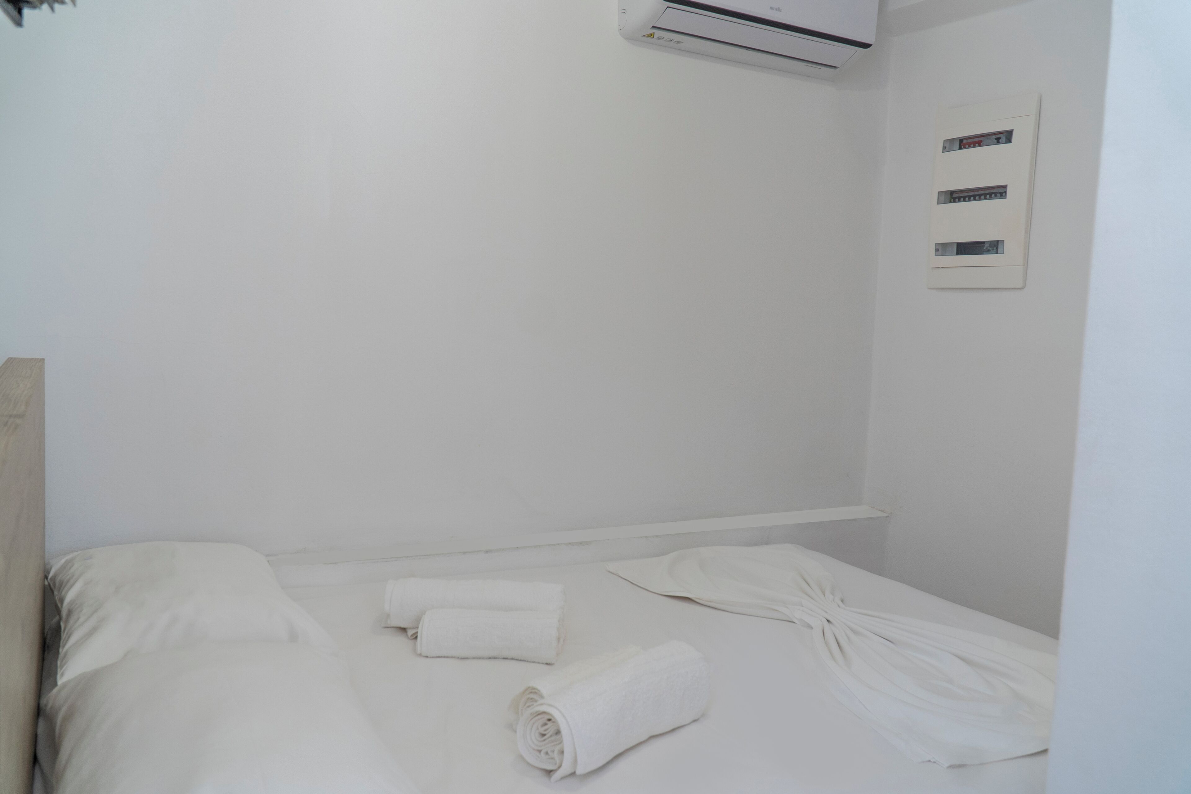 A minimalist hotel room with a bed, two rolled towels, and a wall-mounted air conditioning unit.