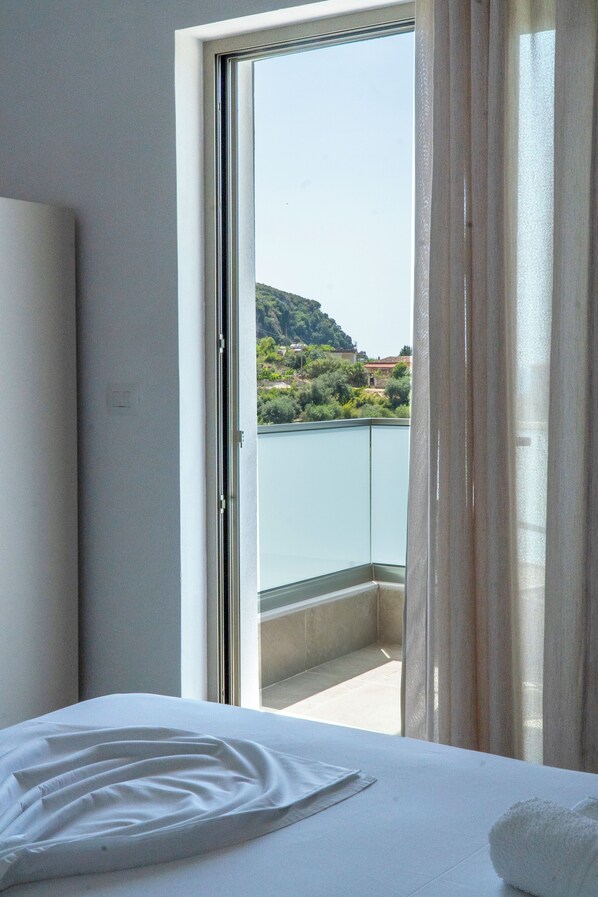 A hotel room with a bed, a window with curtains, and a view of a balcony with greenery and buildings.