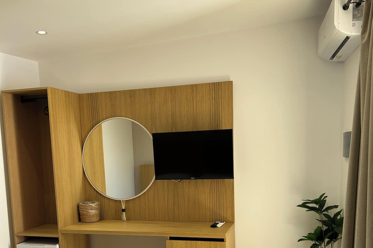 A bedroom with a bed, a wooden desk, a television, a small refrigerator, and a potted plant.