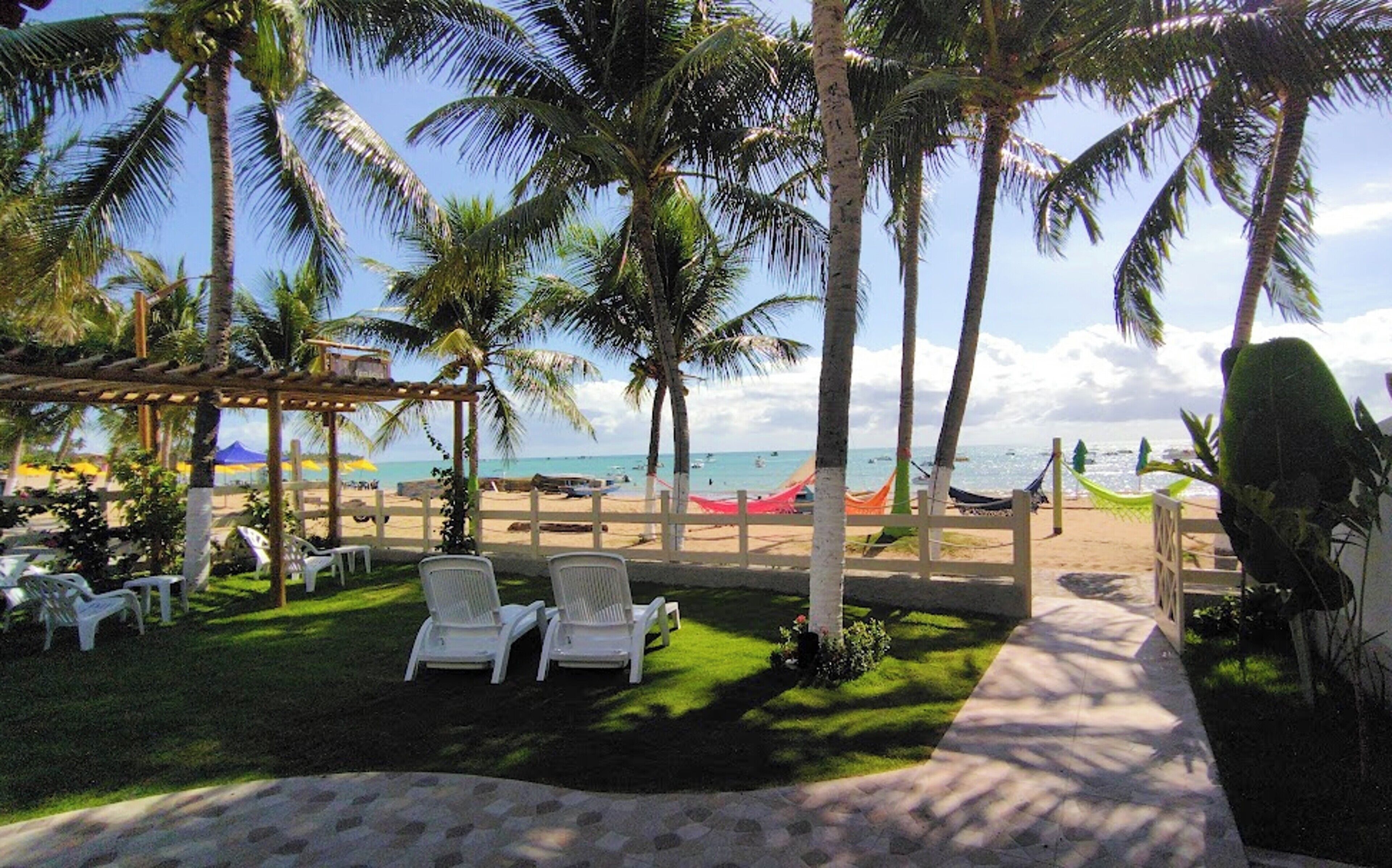 Un patio frente a la playa con sillones blancos, una pérgola y palmeras.
