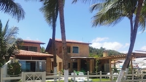 Una casa a due piani in mattoni con tetto in tegole, circondata da un recinto in legno bianco, palme e un cielo azzurro limpido.