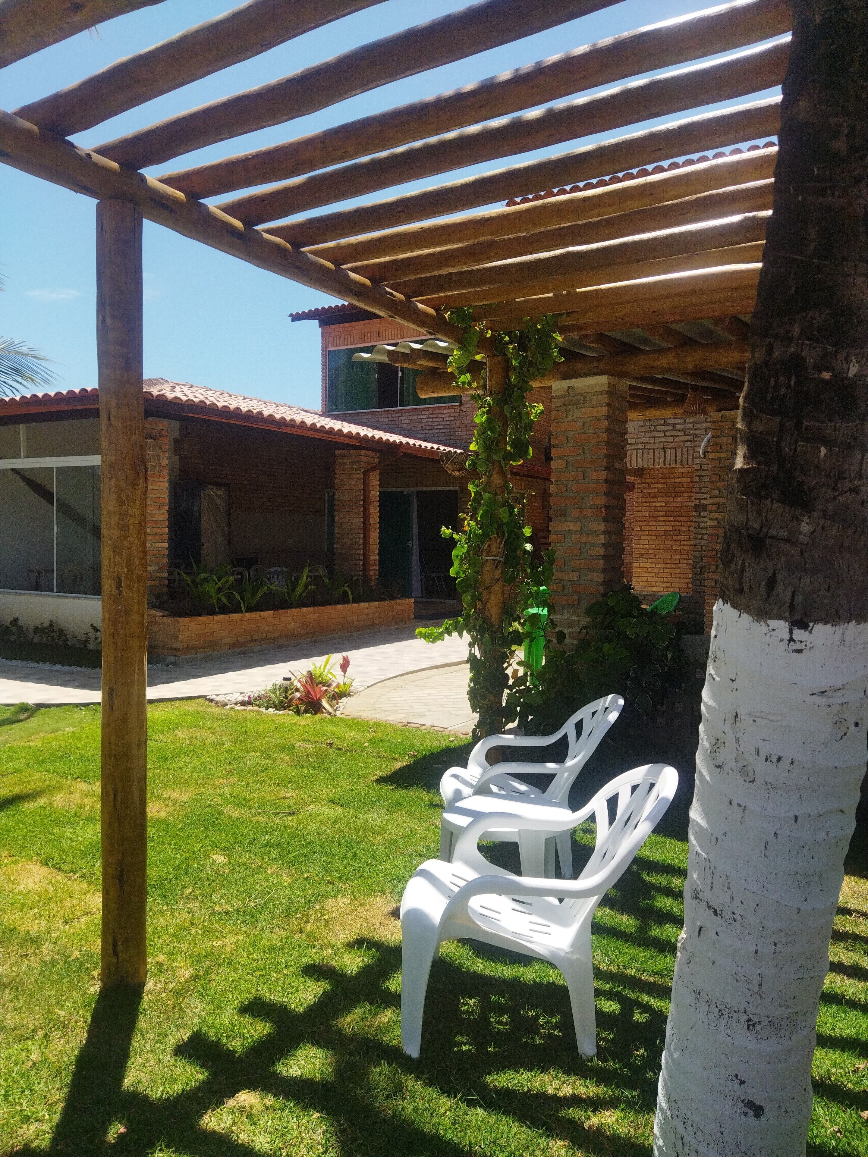A covered patio area with two white plastic chairs, a wooden pergola, and a brick building in the background.