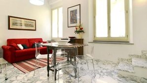 A living room with a red sofa, a dining table with chairs, and a large window with blinds.