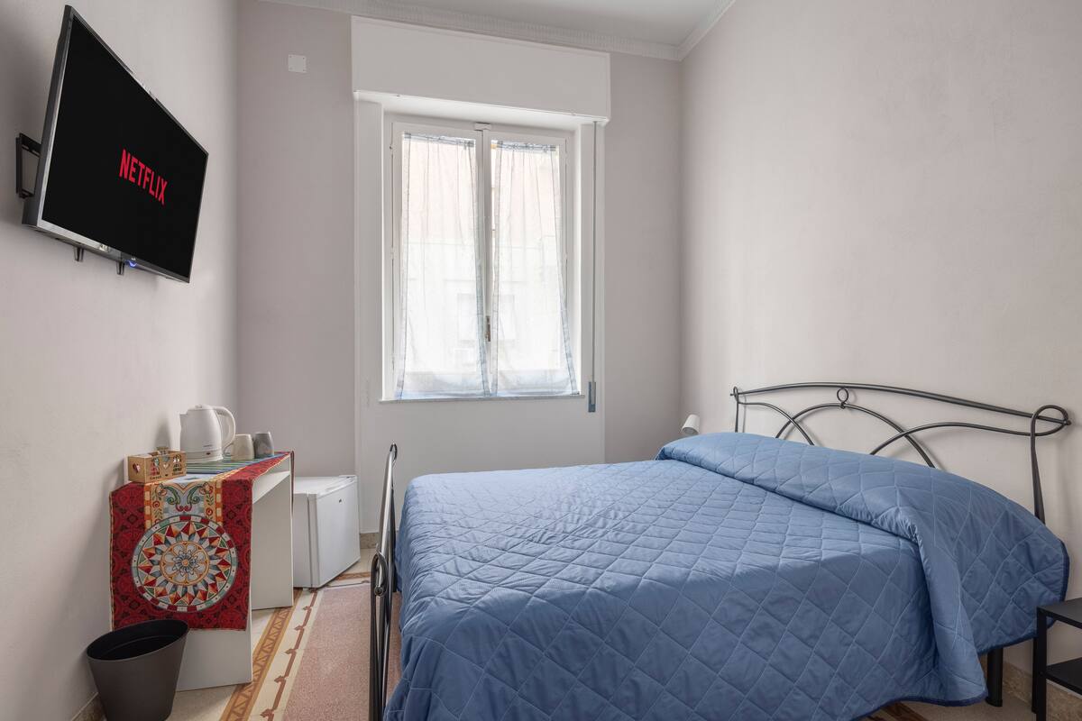 Un dormitorio con cama, televisor montado en la pared, ventana y mesita de noche con tetera y tazas.