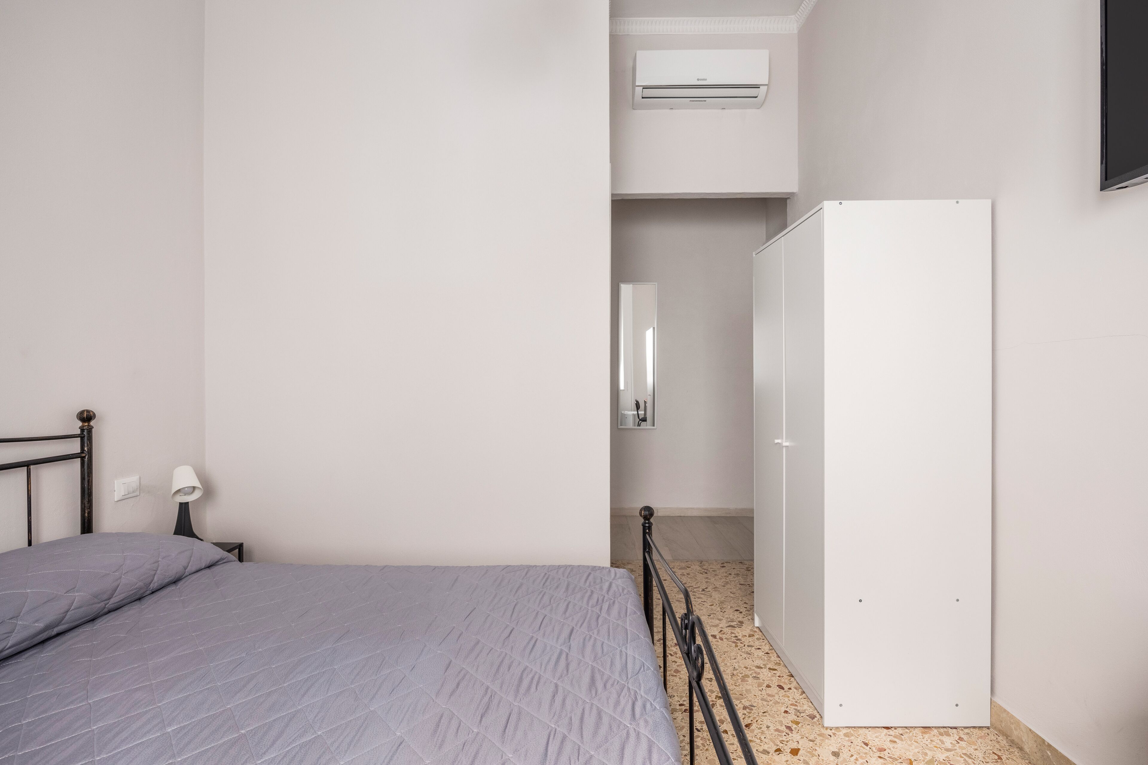 A bedroom with a bed, a nightstand, a lamp, a wardrobe, and an air conditioning unit.