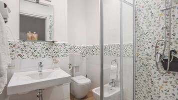 Une salle de bain avec un lavabo, des toilettes et un bidet blancs, une douche avec une porte en verre et des carreaux décorés de motifs floraux.