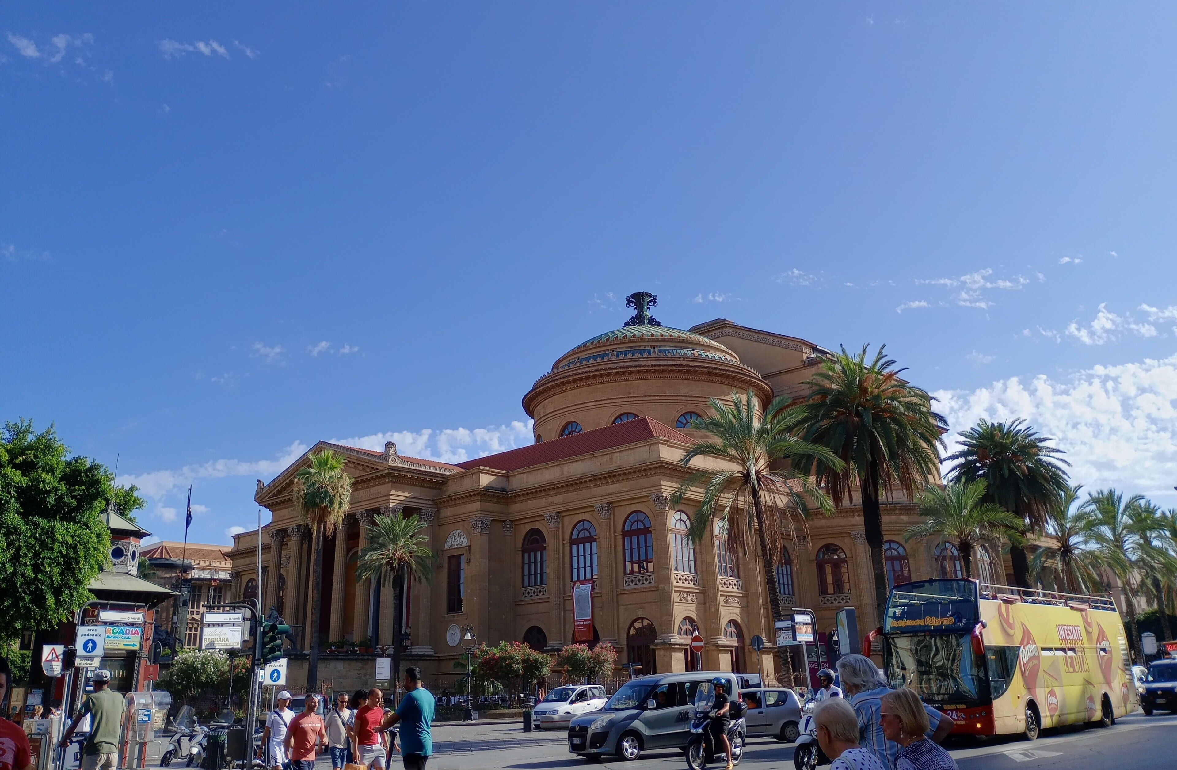 A historic building with classical architecture, including columns and a dome, surrounded by palm trees and busy street activity.