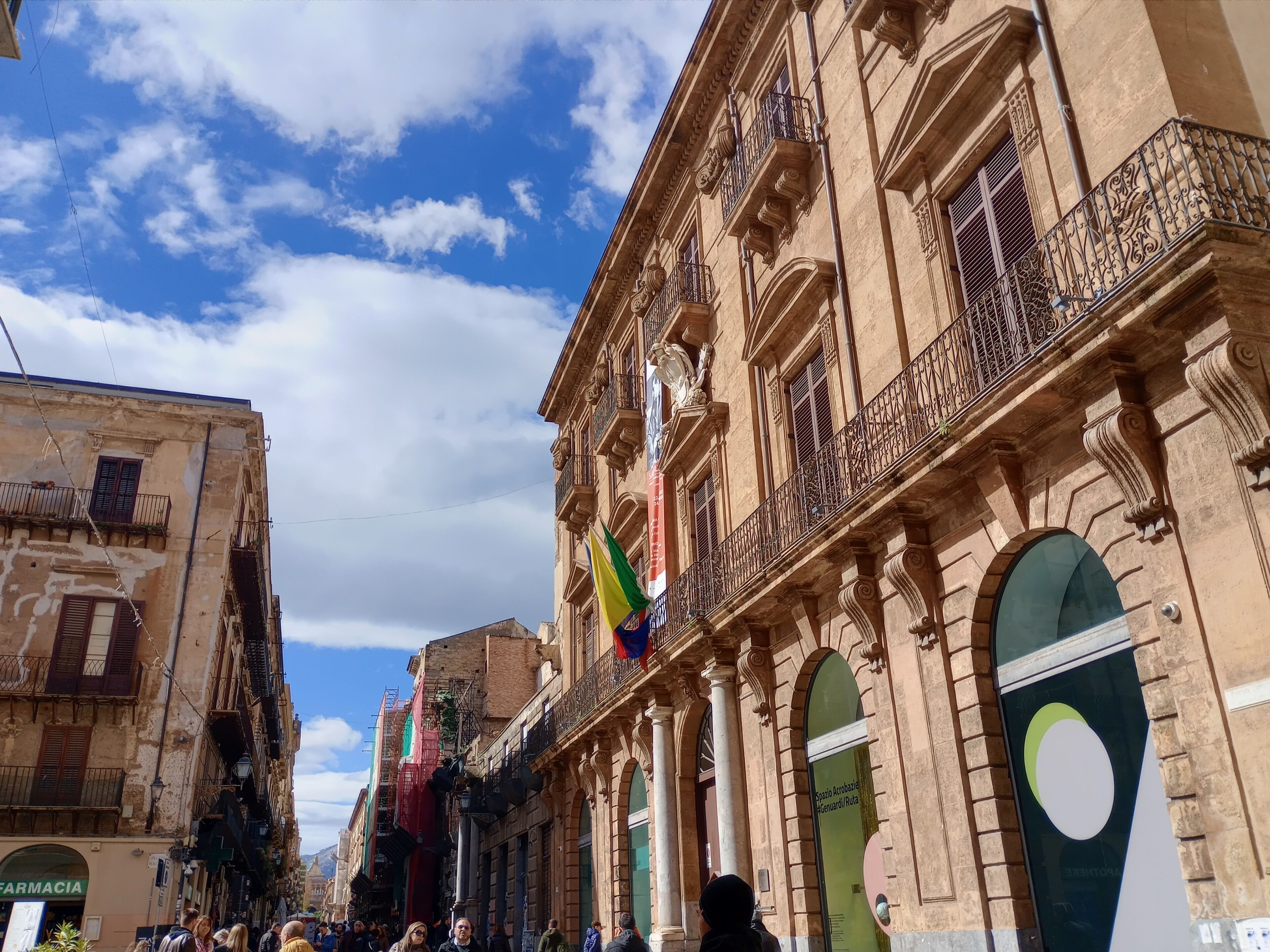 Un edificio histórico con balcones ornamentados y ventanas grandes, un asta con varias banderas, y una calle con peatones.
