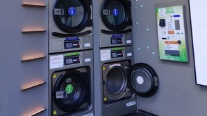 Une laverie équipée de quatre machines à laver et de quatre sèche-linge, chacune étant identifiée par un numéro. Un panneau de commande fixé au mur affiche les instructions et un code QR.
