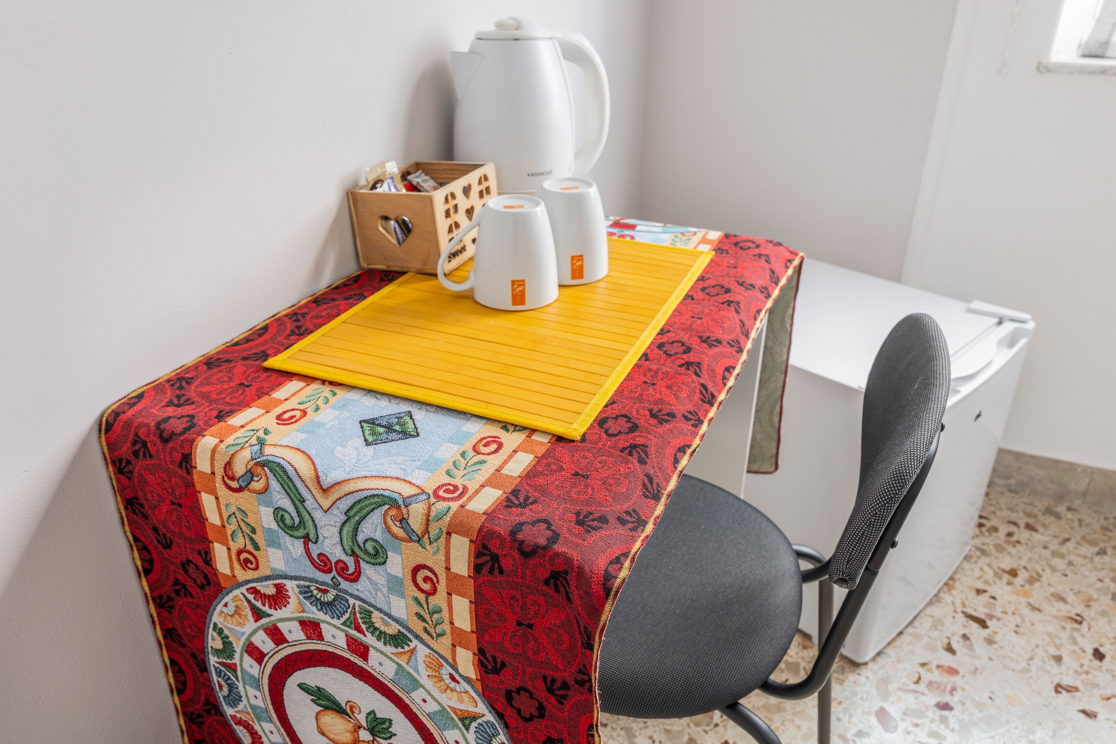 A small, neatly arranged workspace with a patterned tablecloth, a coffee maker, two mugs, and a box of tissues.