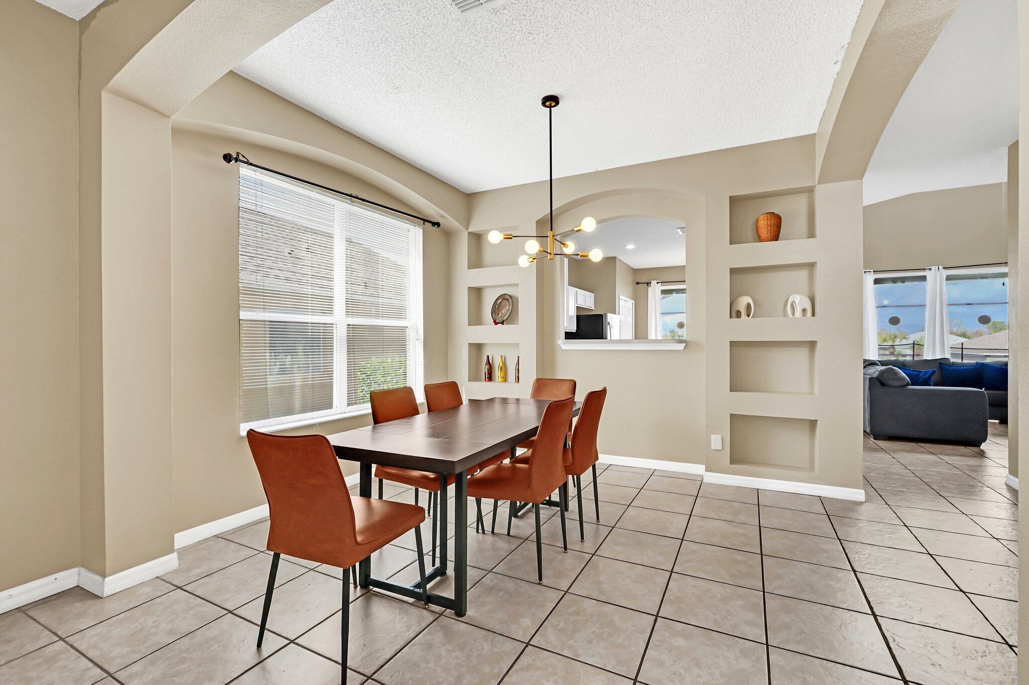 Un coin repas comprenant une table rectangulaire et six chaises, un luminaire au plafond, et donnant sur un salon avec un canapé et une fenêtre.