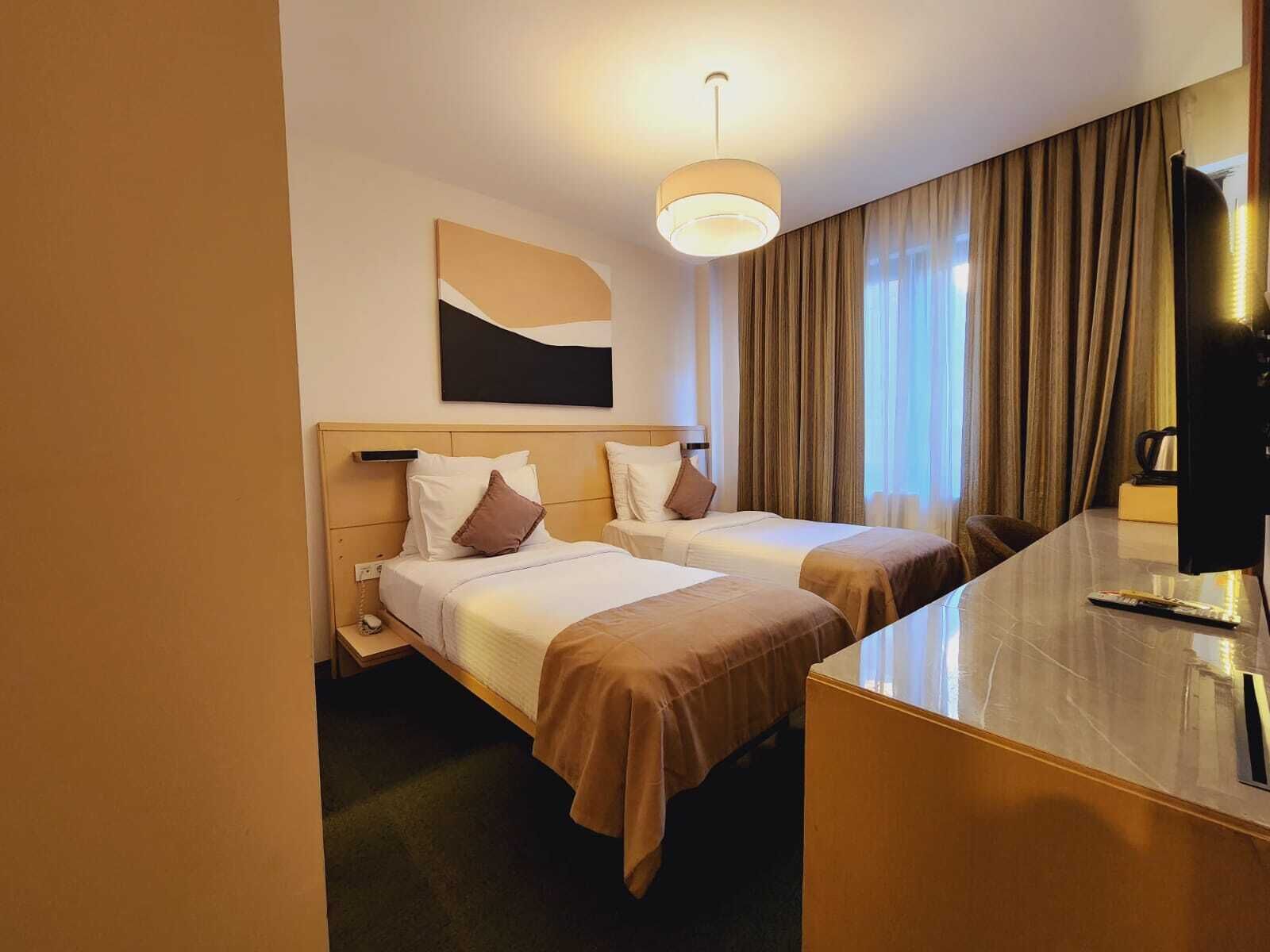 A hotel room with two beds, a desk, a television, and a large window with curtains.