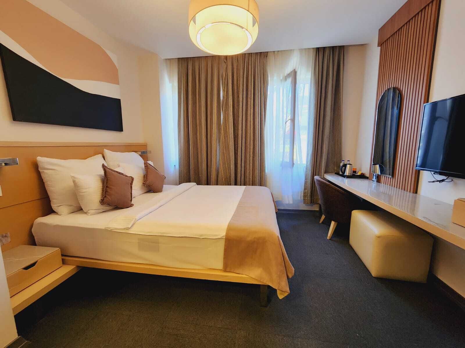 Habitación de hotel con cama, escritorio, silla, televisor y un amplio ventanal con cortinas.