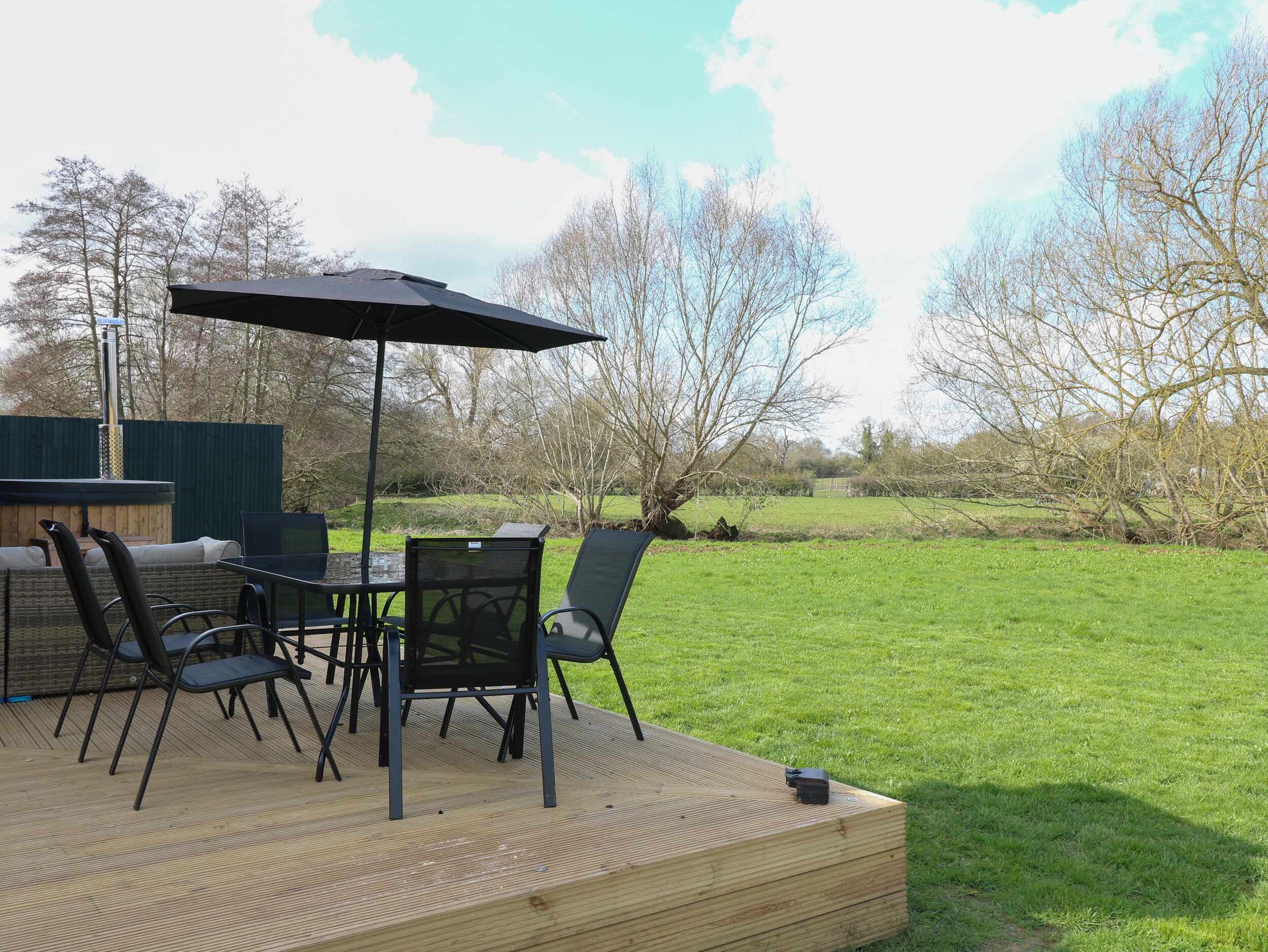 An outdoor deck with a table and chairs, a black umbrella, and a view of a grassy field and trees.