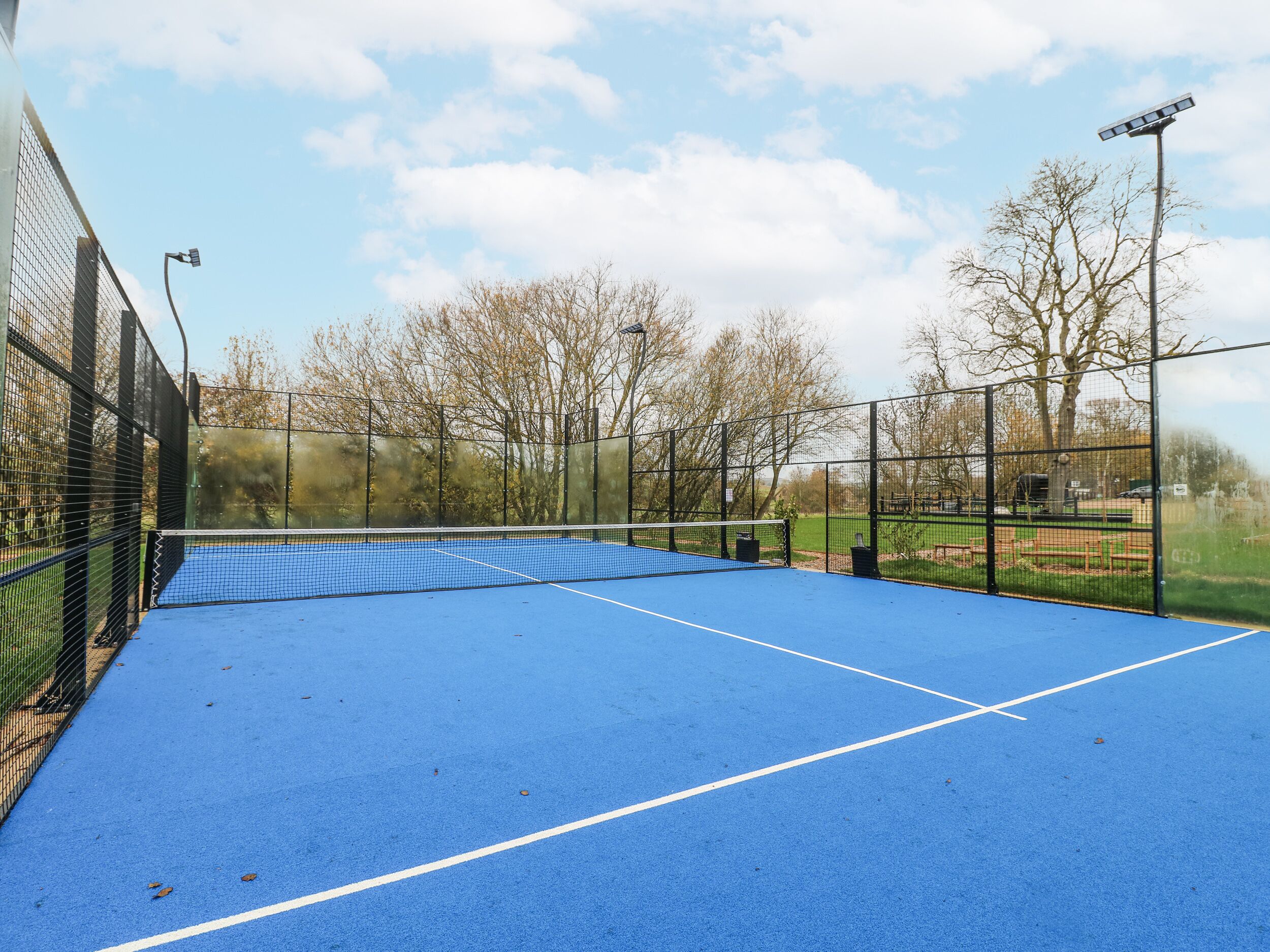 An outdoor tennis court with a blue surface, surrounded by a black fence, and a street lamp nearby.