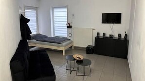 Une petite chambre moderne, équipée d’un lit, d’un canapé noir, d’une petite table et d’un téléviseur fixé au mur.