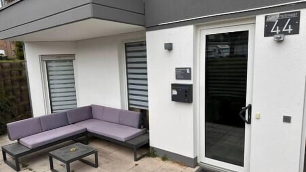 Un espace extérieur moderne aménagé avec un canapé et une petite table, une porte portant un numéro et une boîte aux lettres.