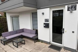 Un espace extérieur moderne aménagé avec un canapé et une petite table, une porte portant un numéro et une boîte aux lettres.