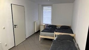 Une petite pièce, peu meublée, avec un lit simple, un radiateur, une fenêtre avec des stores et une porte.