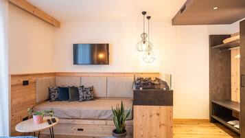 A modern living room with a wooden floor, a sofa with patterned cushions, a mounted TV, and a hanging light fixture.