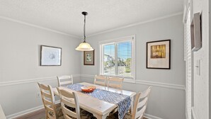 A dining room with a wooden table, chairs, and a chandelier. There are framed pictures on the walls and a window with blinds. - Waipani D13 - Beach is Just Across the Street (North Myrtle Beach)