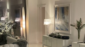 A modern living room with a white sofa, fluffy grey pillows, a round coffee table with three wine glasses, a decorative plant, and a painting on the wall.