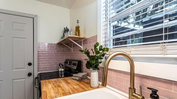 A kitchen with a wooden countertop, a white sink with a gold faucet, a window with blinds, and a brick backsplash. - Family & Dog Friendly Fun New Renovation w/ Pool!
 (Savannah)