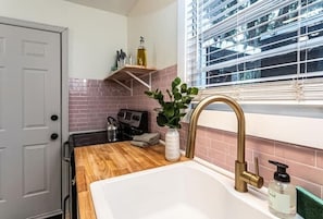 A kitchen with a wooden countertop, a white sink with a gold faucet, a window with blinds, and a brick backsplash. - Family & Dog Friendly Fun New Renovation w/ Pool!
 (Savannah)