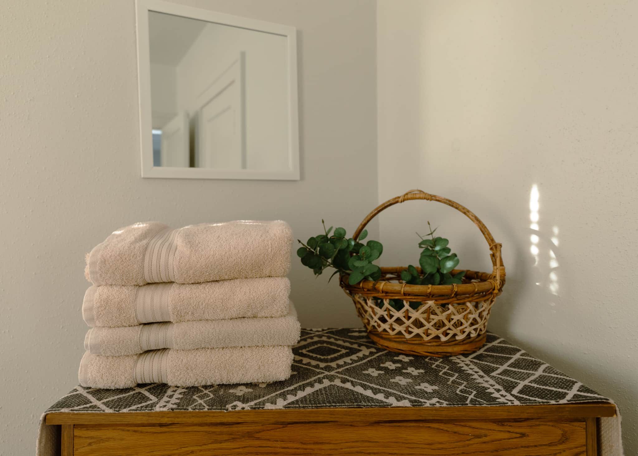 A stack of towels on a wooden table with a patterned rug, a wicker basket with greenery, and a mirror on the wall.