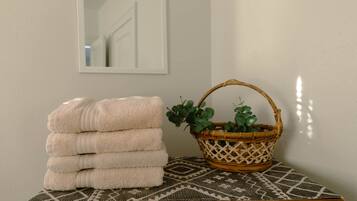 A stack of towels on a wooden table with a patterned rug, a wicker basket with greenery, and a mirror on the wall.