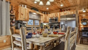 Interior de una cabaña de madera con una mesa de comedor central, sillas de madera, una cocina con electrodomésticos modernos y una chimenea acogedora.