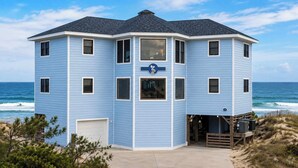 A blue, multi-story building with a large bay window and a garage, located near the beach with ocean views. - Pompous Pelican - Luxury Oceanfront Escape 6 BR/6B (Nags Head)
