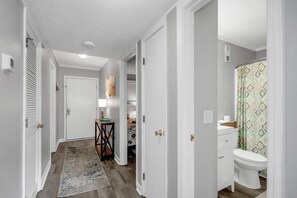 A hallway with a wooden floor, a rug, a desk with a lamp, and a bathroom with a toilet and patterned shower curtain.