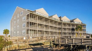 A multi-story building with a balcony on the second floor, a wooden deck in front, and a clear sky. - Seawatch Condo | Marsh View & Pool  | Cozy 2BR  |  Relaxing Amenities Free Travel Insurance on weekly rentals (Murrells Inlet)