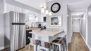 A modern kitchen with a stainless steel refrigerator, a granite countertop, bar stools, a clock, and a framed picture on the wall.