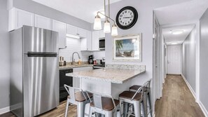 A modern kitchen with a stainless steel refrigerator, a granite countertop, bar stools, a clock, and a framed picture on the wall.
