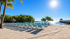 A beach with rows of sun loungers, a palm tree, and a thatched-roof hut. - Key Escape | Ocean Pointe 3415 | Oceanfront 2BR Condo in Key Largo (Tavernier)
