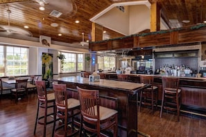A spacious bar area with wooden bar stools and a long counter, surrounded by large windows and a high ceiling with exposed beams.