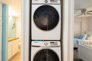 Une machine à laver et un sèche-linge superposés dans une pièce qui contient également un lit et un ventilateur de plafond.